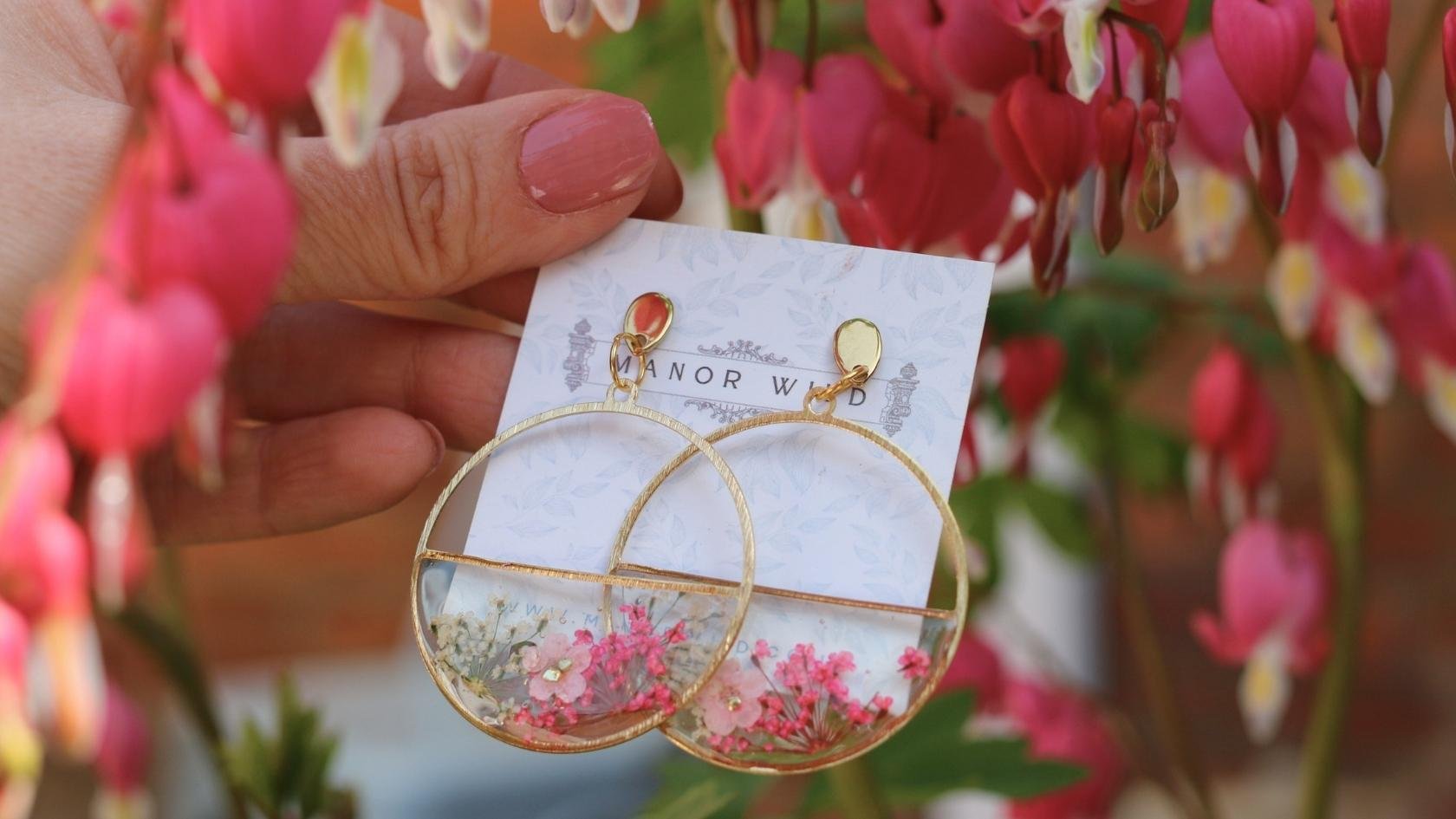 A pair of gold hoop earrings with watercolor flower designs, held against a backdrop of pink and white flowers.