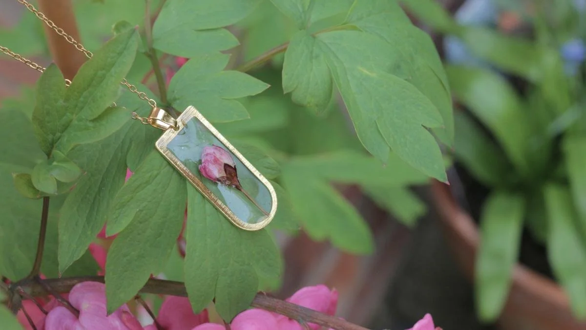 A gold necklace with a rectangular pendant, featuring a pressed pink rose, hanging on green leaves.