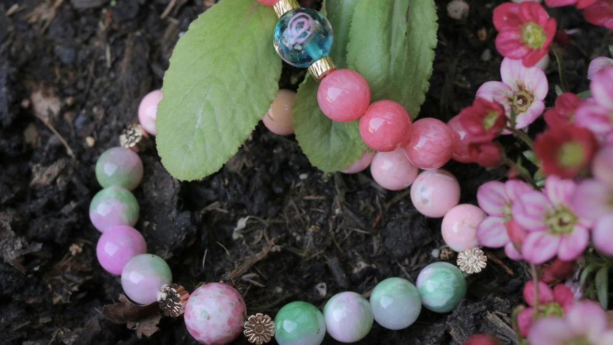 A pastel-colored bead bracelet resting on soil among green leaves and pink flowers.