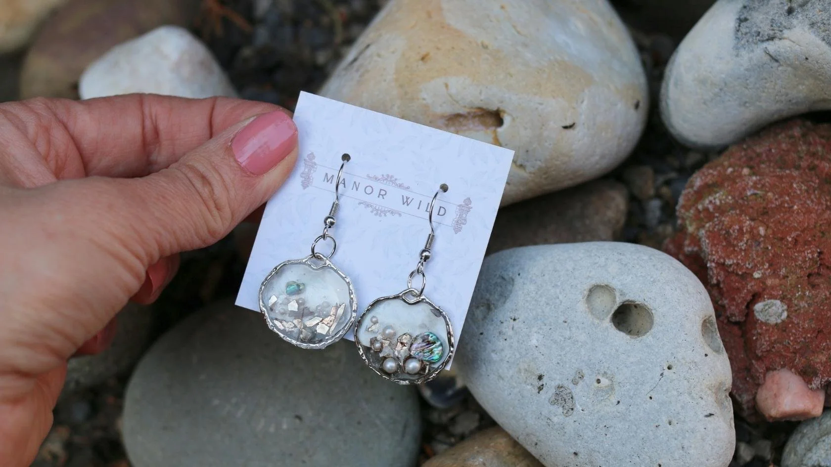 A hand holding a pair of dangling earrings on a white card with the text 'Manor Wild,' surrounded by various rocks on the ground.