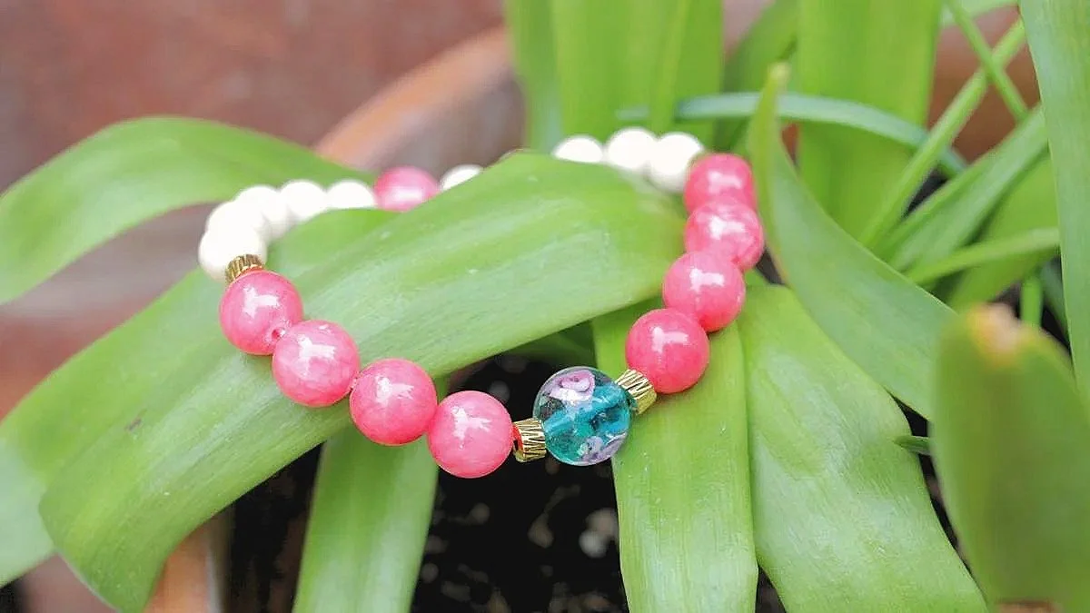 A beaded bracelet with pink, white, and blue beads resting on green leaves of a potted plant.