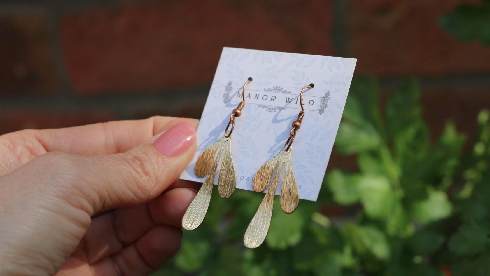 Hand holding a pair of gold and rose gold drop earrings with curved, elongated shapes on a white card with the brand name 'Manor Wild', against a green leafy background.