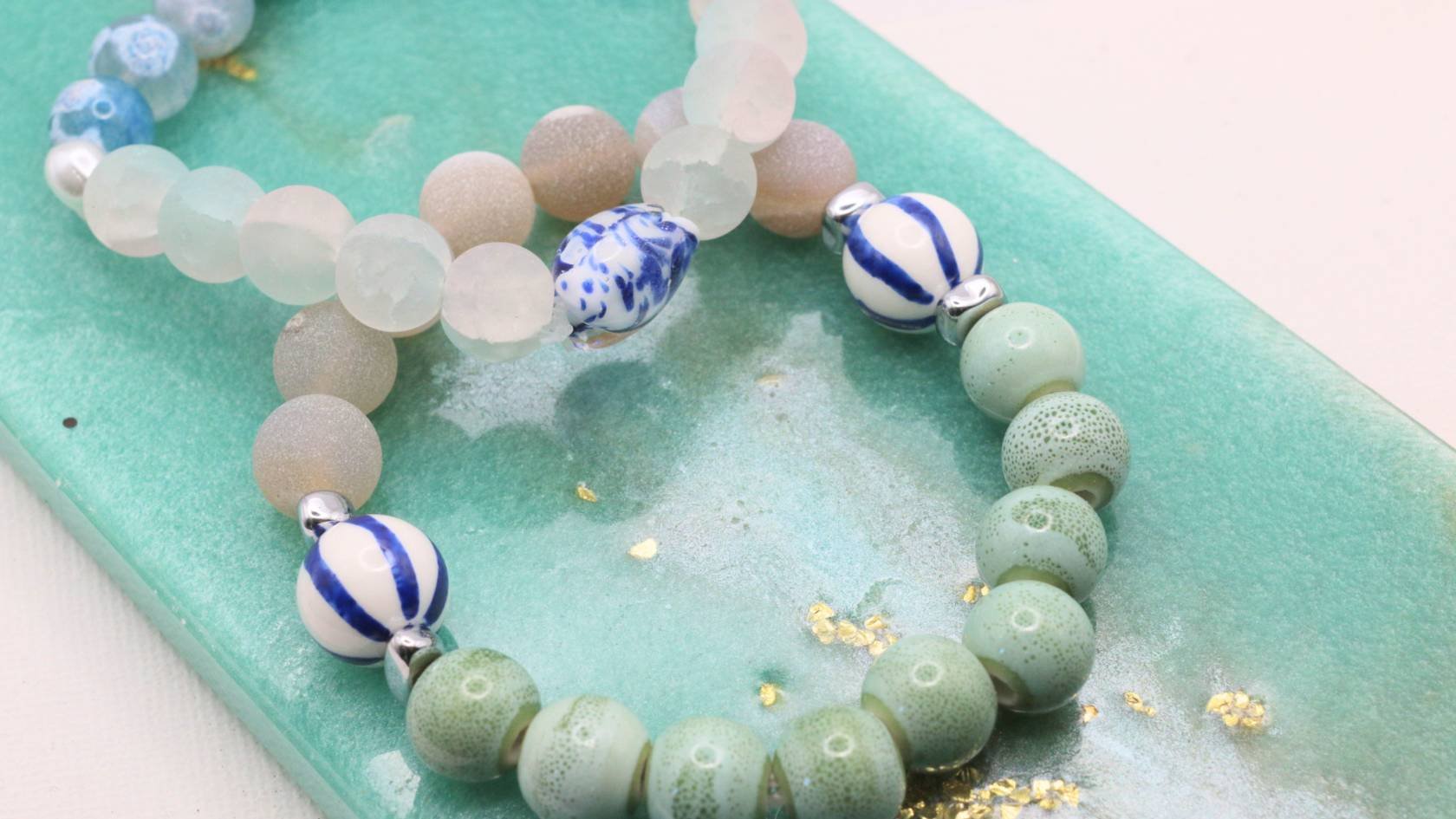 Close-up of a beaded bracelet with pastel green, white, and blue beads, placed on a green surface with gold flecks.