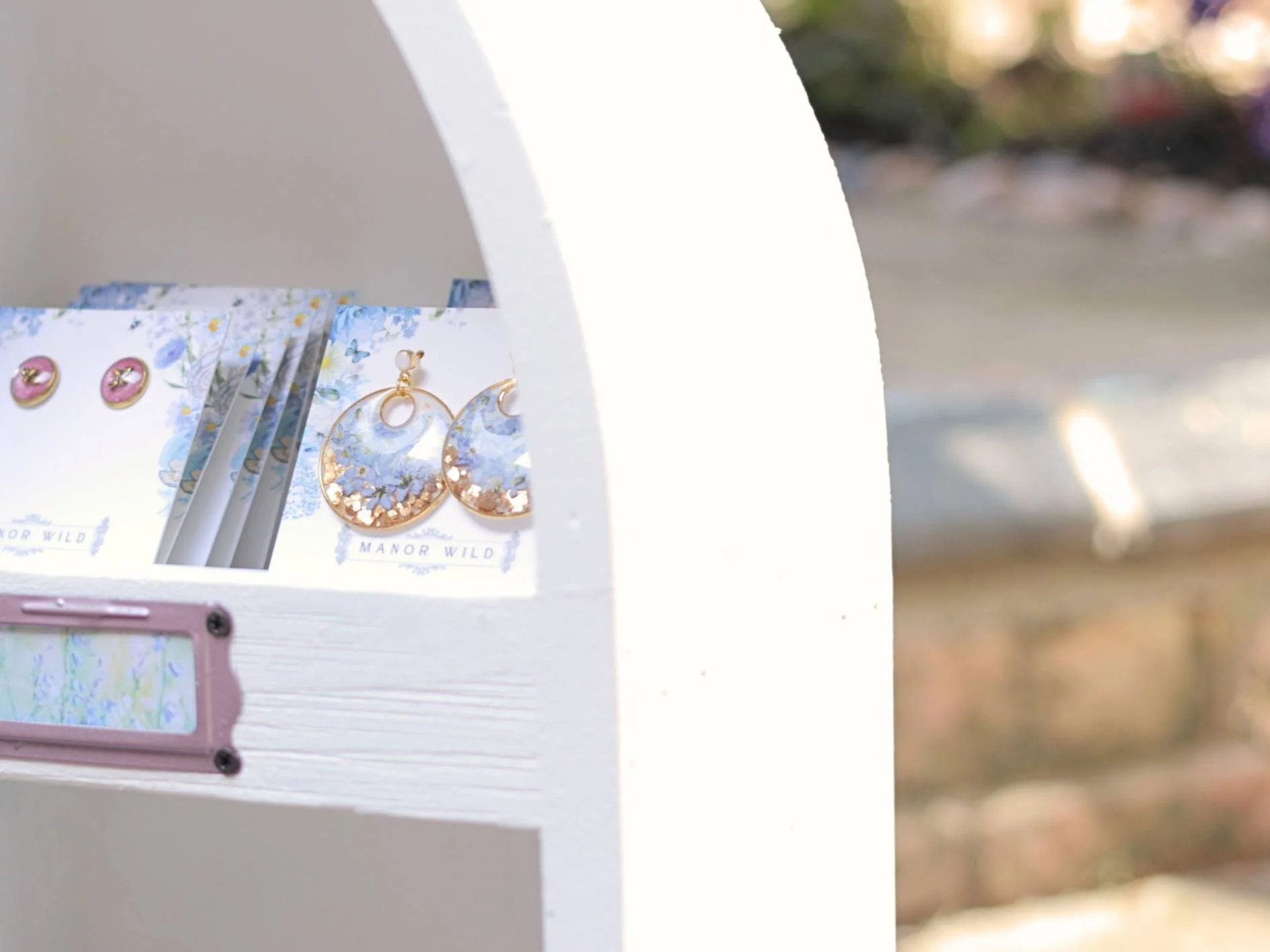 A white jewelry display stand featuring gold and floral earrings next to a floral-themed jewelry box. The background is blurred with natural outdoor lighting.
