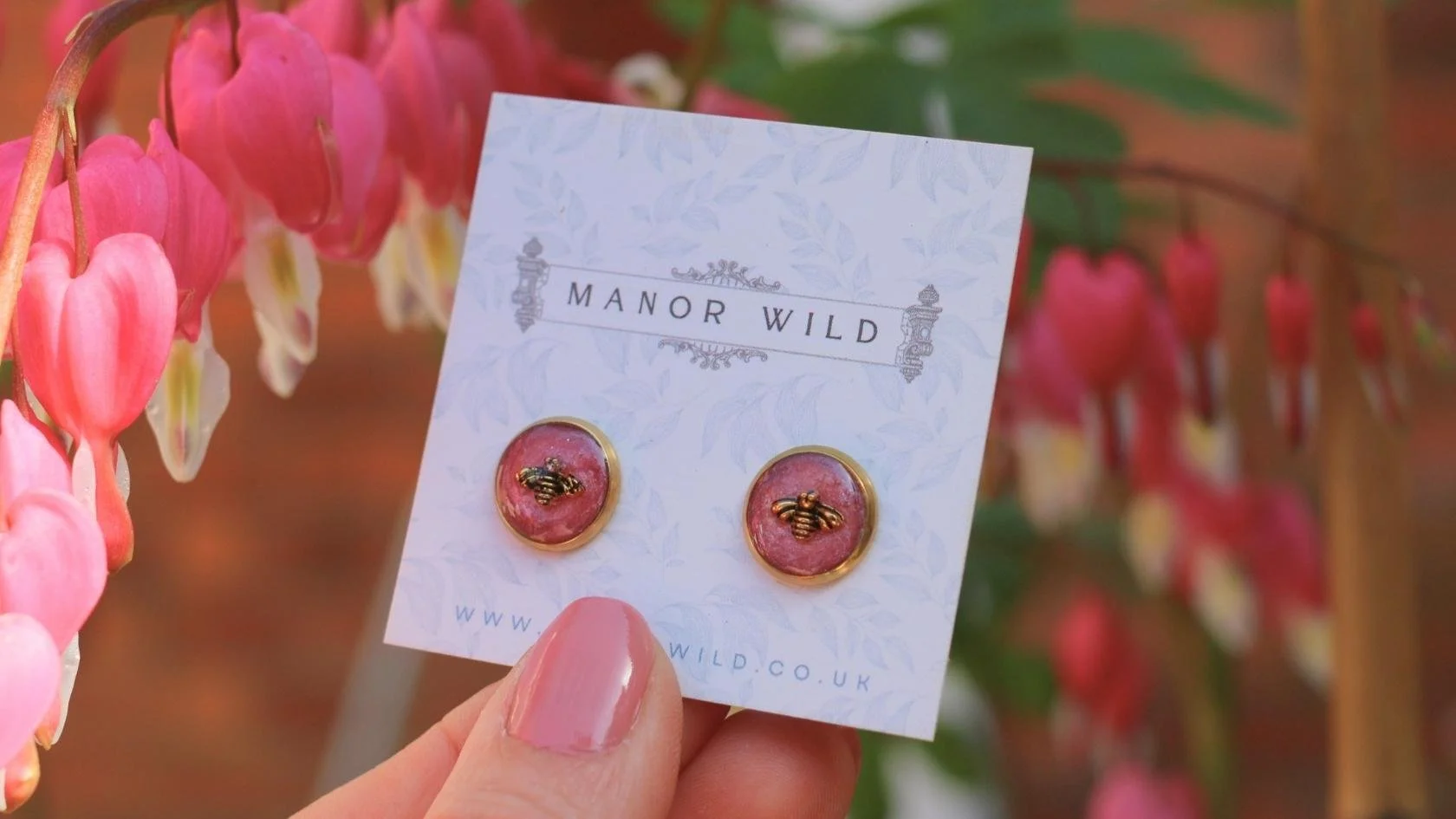 Pink earrings with bee designs displayed on a white card from Manor Wild, held by a person with light pink painted nails, with pink bleeding heart flowers in the background.