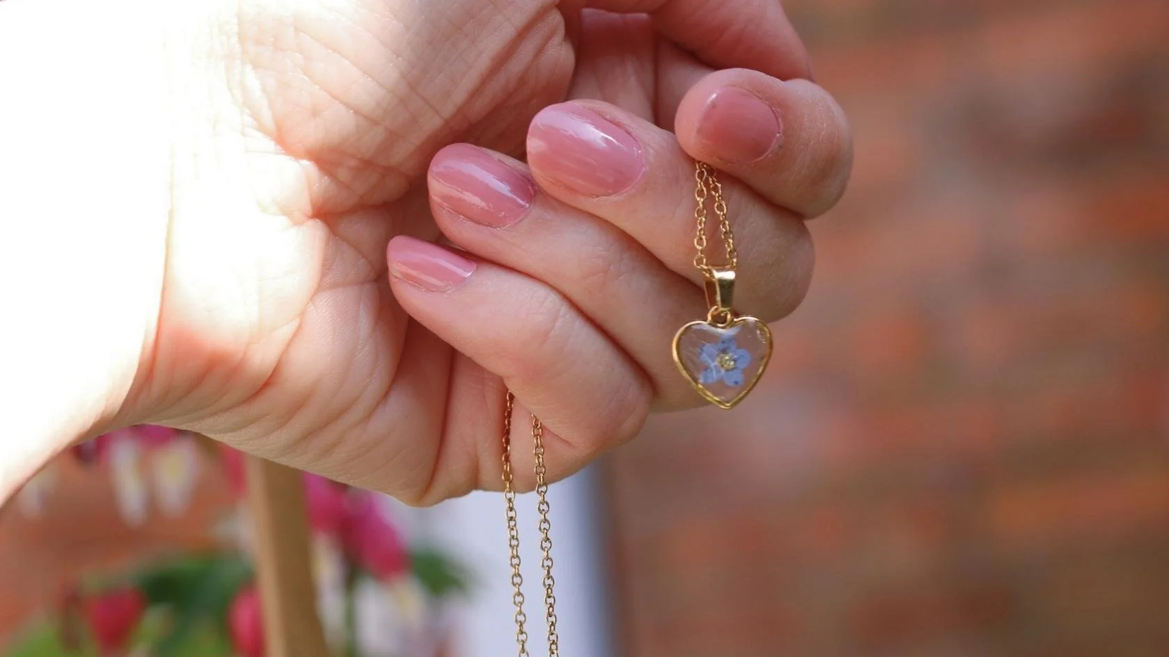 A hand holding a gold necklace with a heart-shaped pendant containing a blue flower with a yellow center.