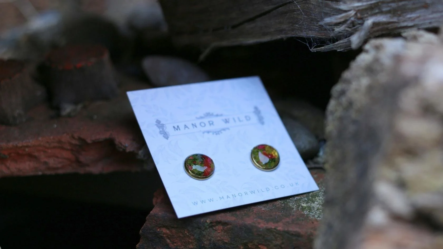 A pair of round earrings with red and green Christmas-themed wreath designs on a white card from Manor Wild, placed among rocks.