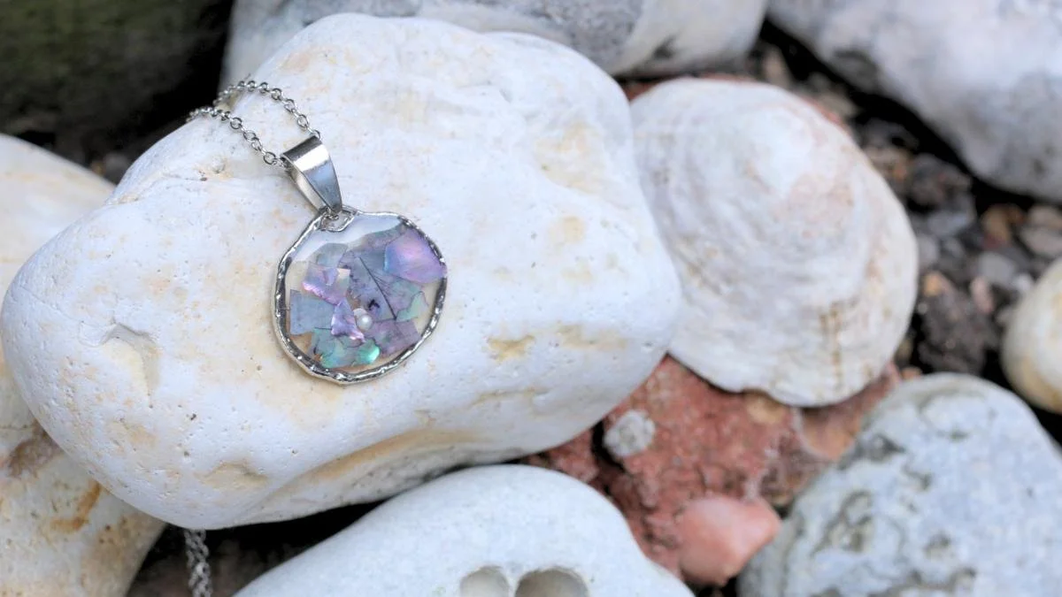 A silver necklace with a circular pendant containing iridescent, broken shell pieces, resting on white rocks.