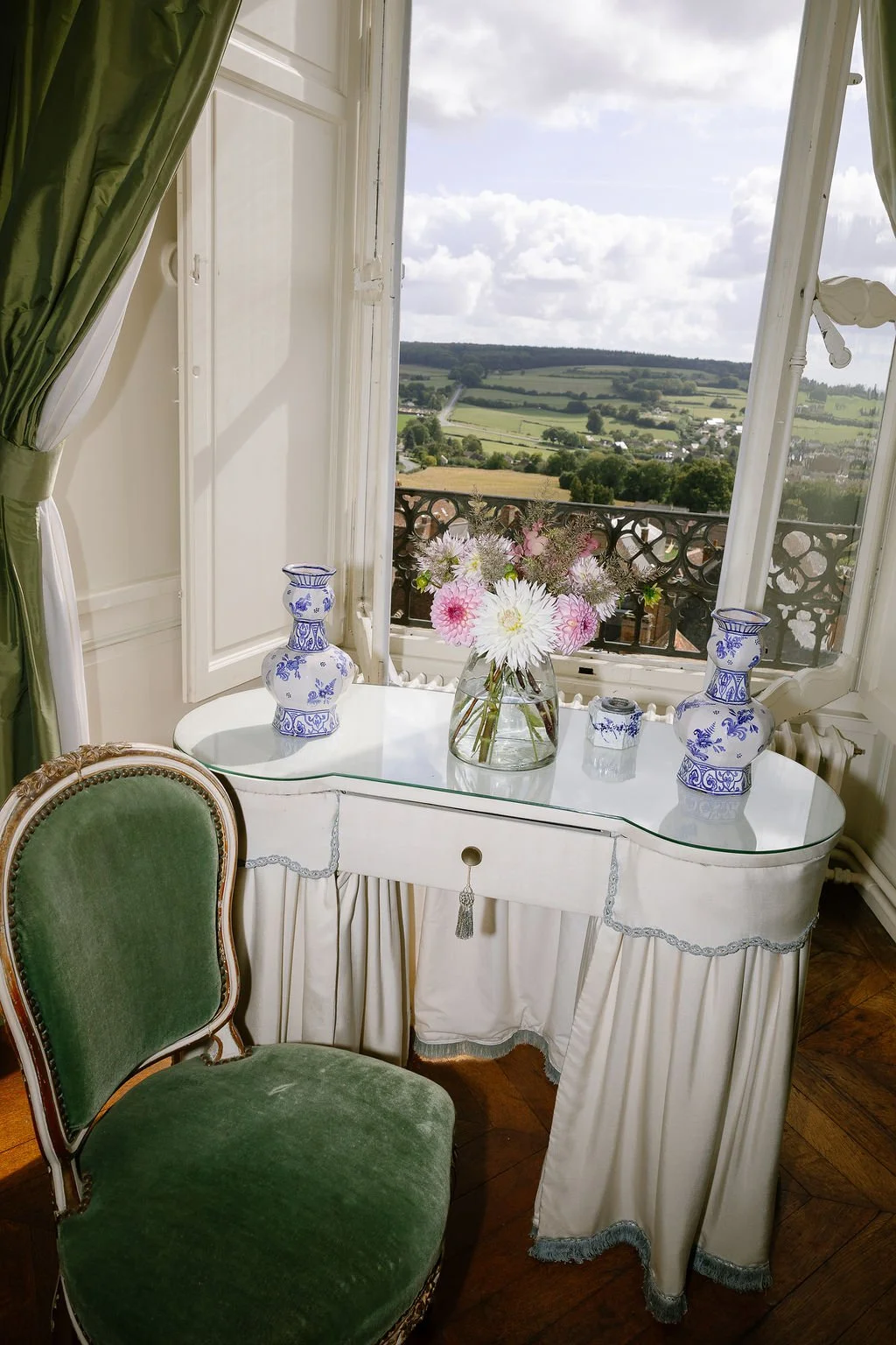 A vintage green velvet chair beside a white antique vanity table with a glass top, decorated with blue and white porcelain vases, a glass jar, and a flower bouquet. An open window shows a scenic countryside landscape with fields and a cloudy sky.