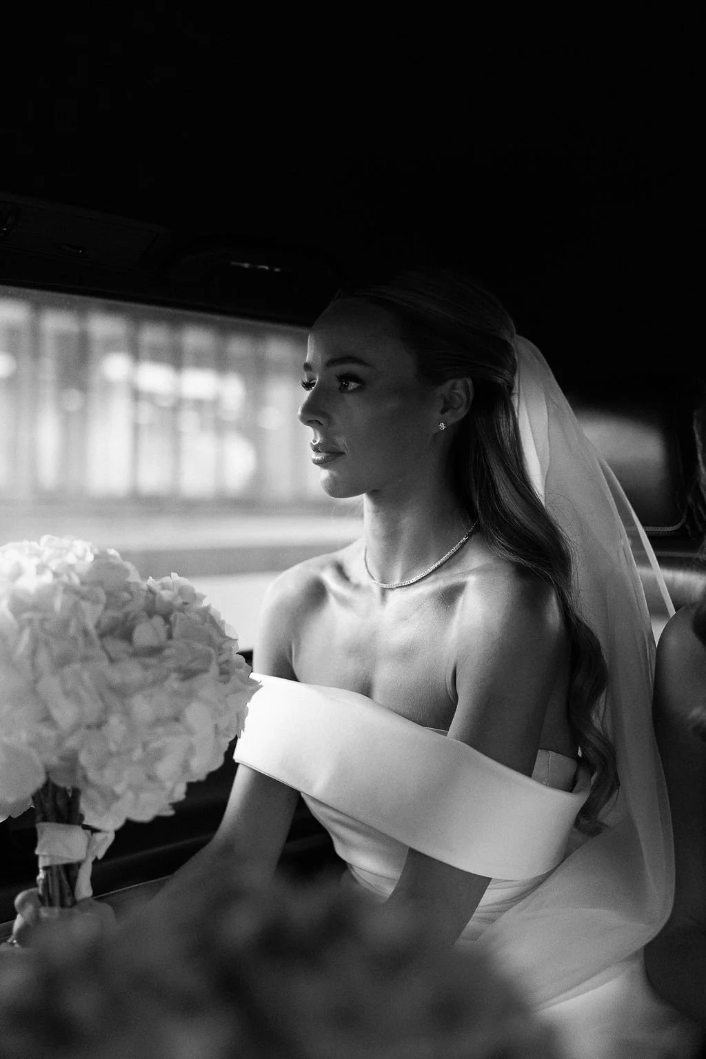 Black and white photo of a woman in a wedding dress sitting inside a car, holding a bouquet of flowers, with a serious expression.