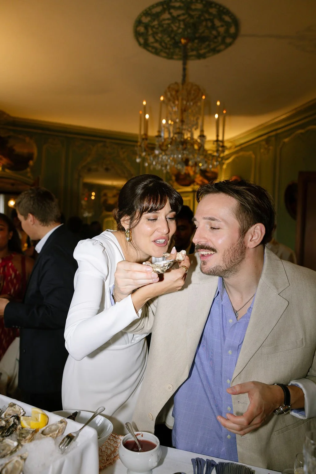 A woman feeds a man oysters at a formal dinner party in an ornate room with a chandelier.
