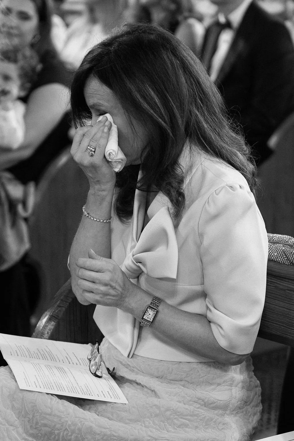 A woman with dark hair, crying and wiping her eyes with a tissue during a formal event, wearing a light-colored blouse with a bow, sitting at a table with papers and reading glasses.