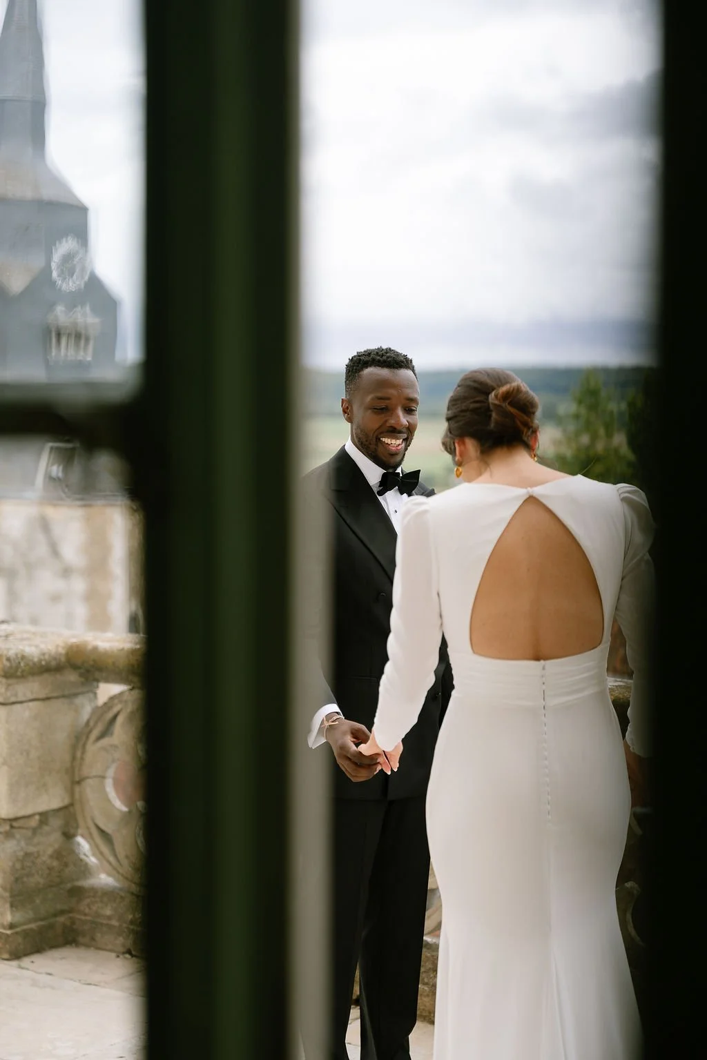 A bride and groom holding hands during their wedding ceremony outside, she wearing a white dress with an open back, he in a black tuxedo and bow tie, with a scenic outdoor backdrop.