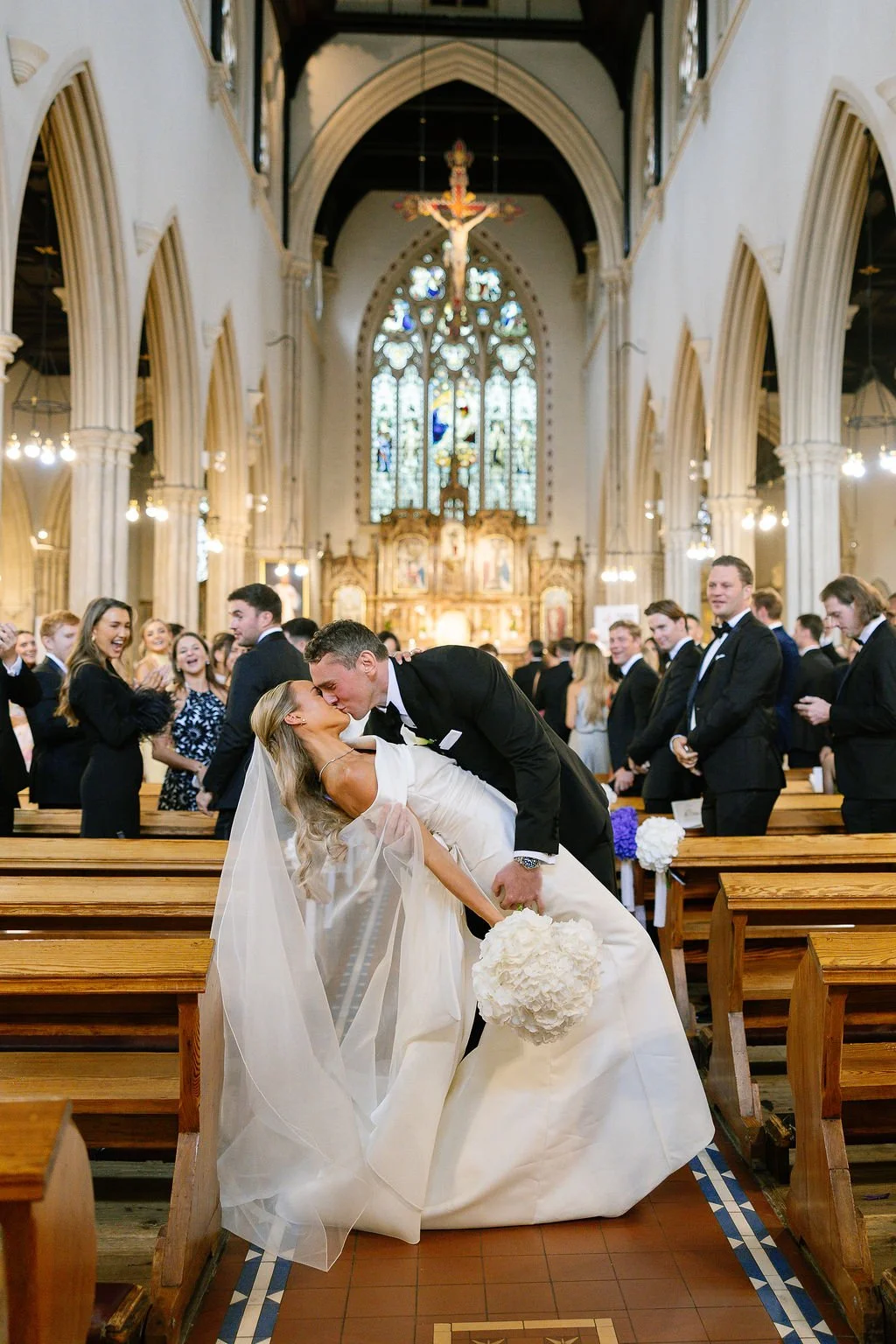 A bride and groom kiss in a church with arching columns and stained glass windows while wedding guests stand and smile in the background.
