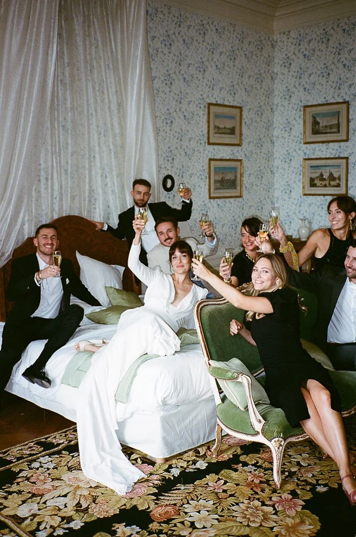 A group of friends celebrating in a hotel room, toasting with champagne while sitting and standing around a bed and vintage green chair.