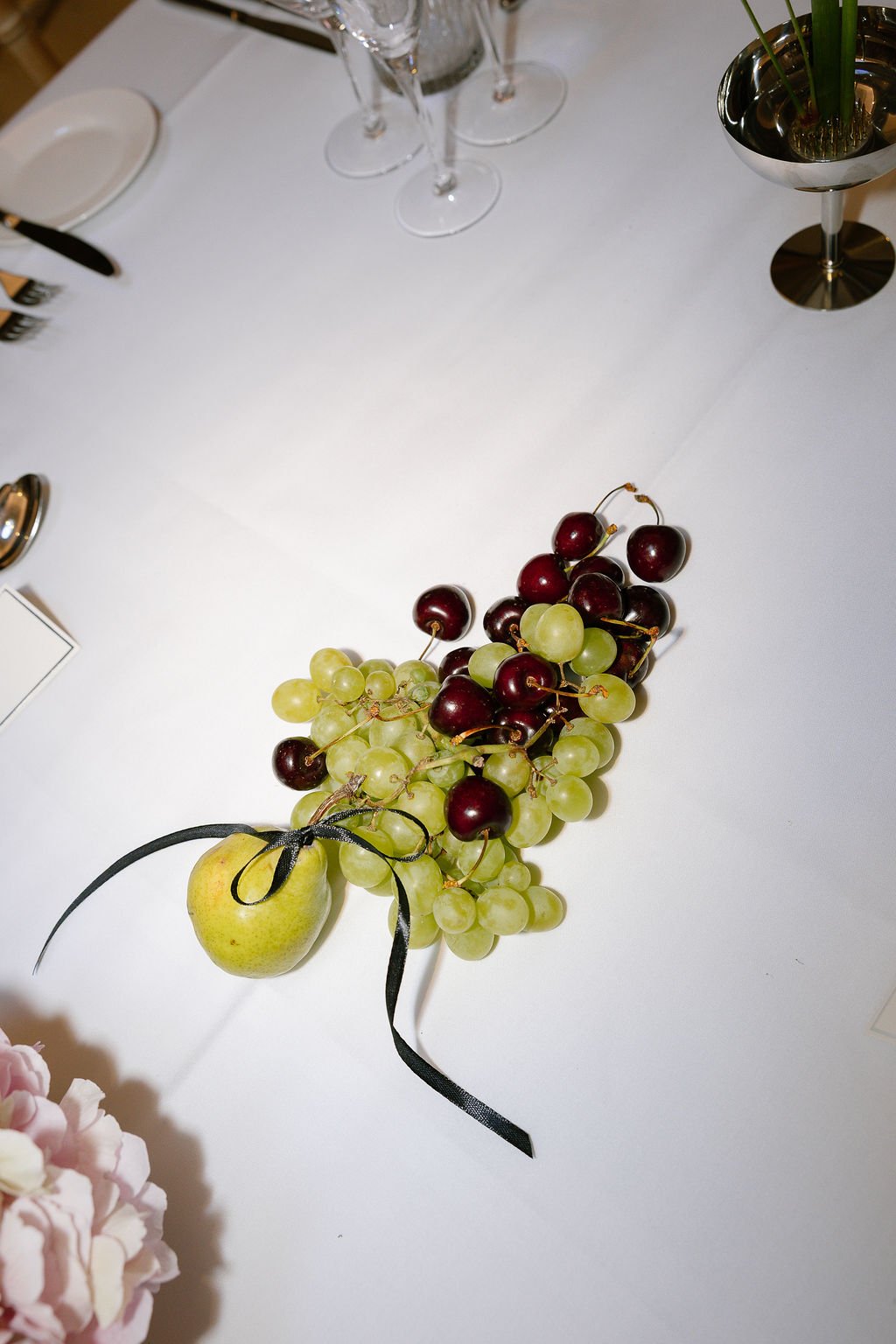 A decorative arrangement of green and red grapes with a small decorative apple tied with a black ribbon, placed on a white tablecloth with glassware and a flower in the background.