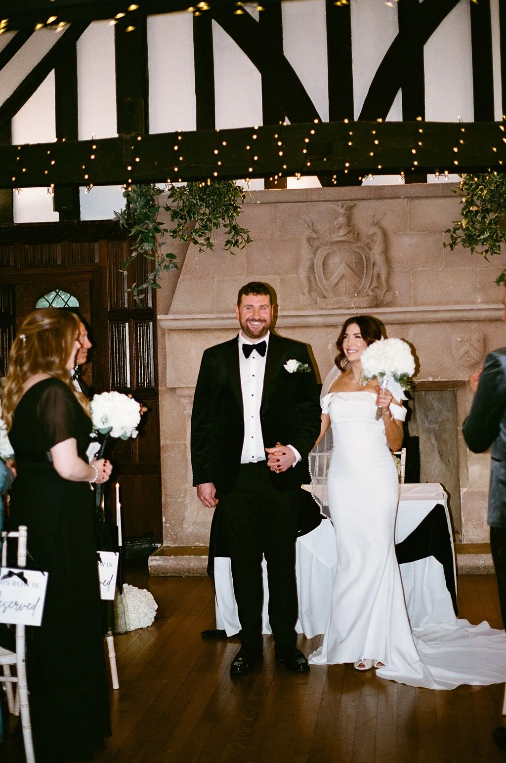 A bride and groom standing together at their wedding ceremony inside a decorated venue, with guests around them and holding bouquets of flowers.