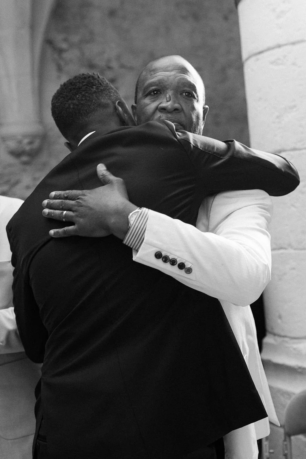 Two men embrace, one is an older man in a white jacket and the other is a younger man in a dark jacket, in a heartfelt hug.