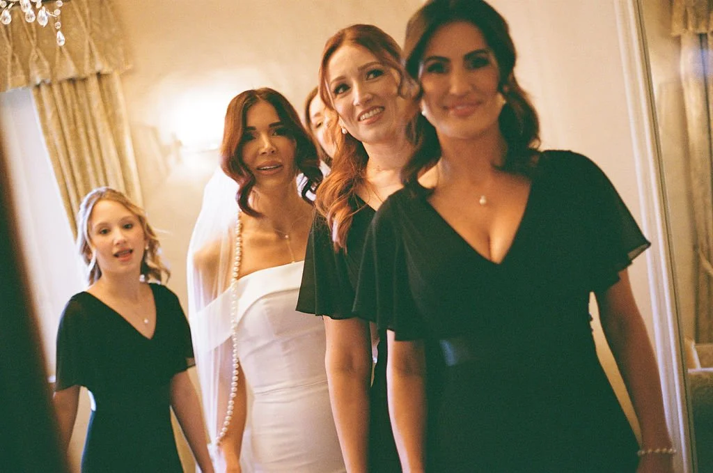 Group of women in formal attire, smiling, standing indoors near a mirror.