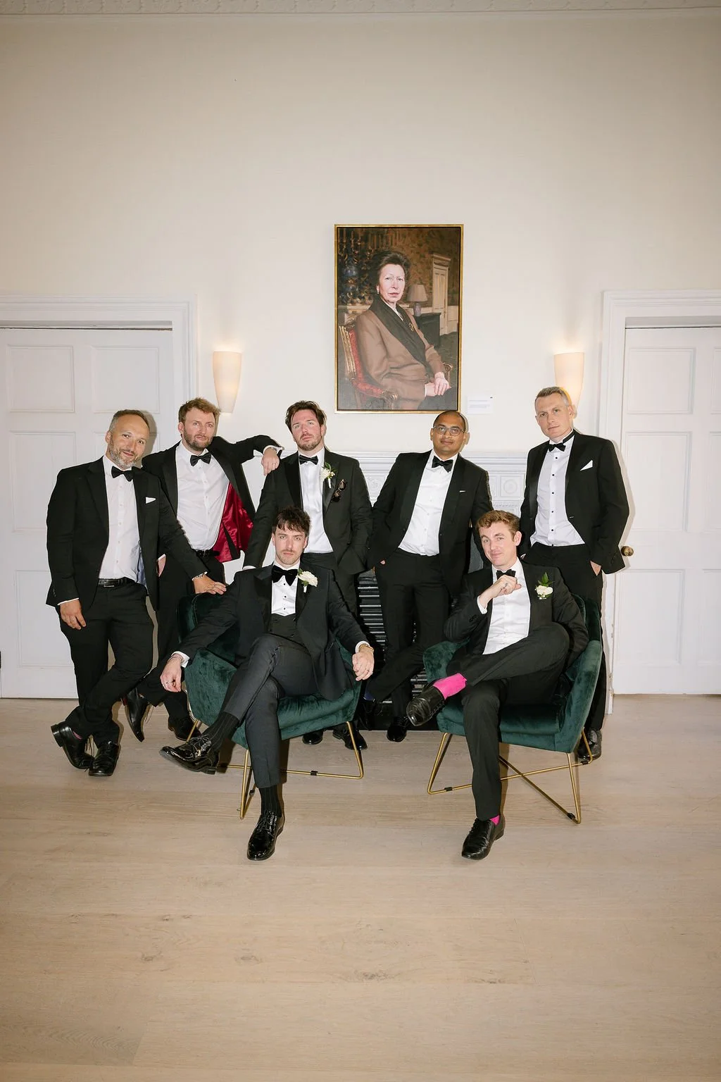 A group of seven men in tuxedos posing in a formal room with white walls and a portrait of a woman hanging on the wall.