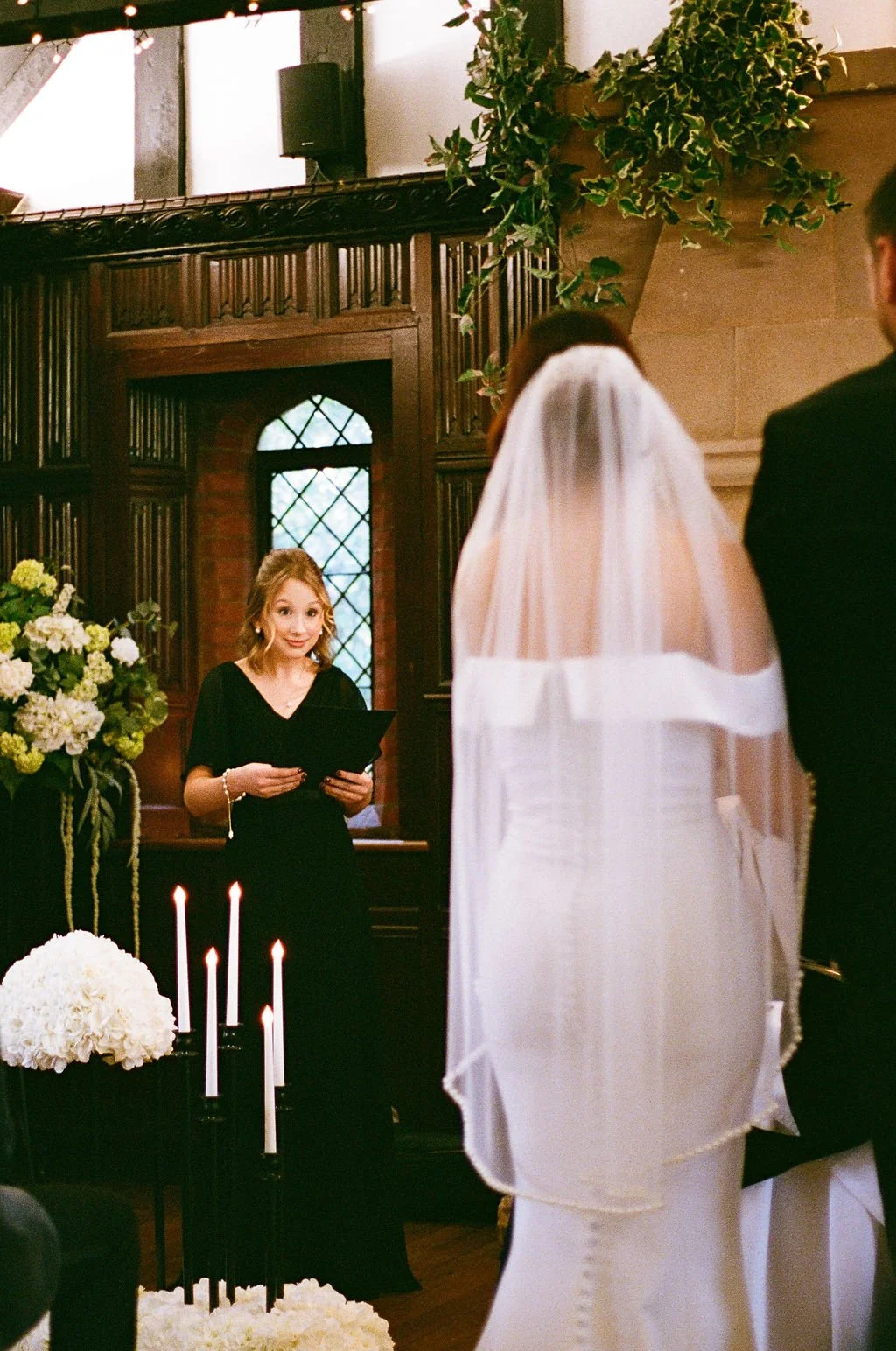 A wedding ceremony with a officiant reading vows or a speech, a bride with a white veil and gown, and a groom in a black suit, in an indoor venue decorated with flowers and candles.