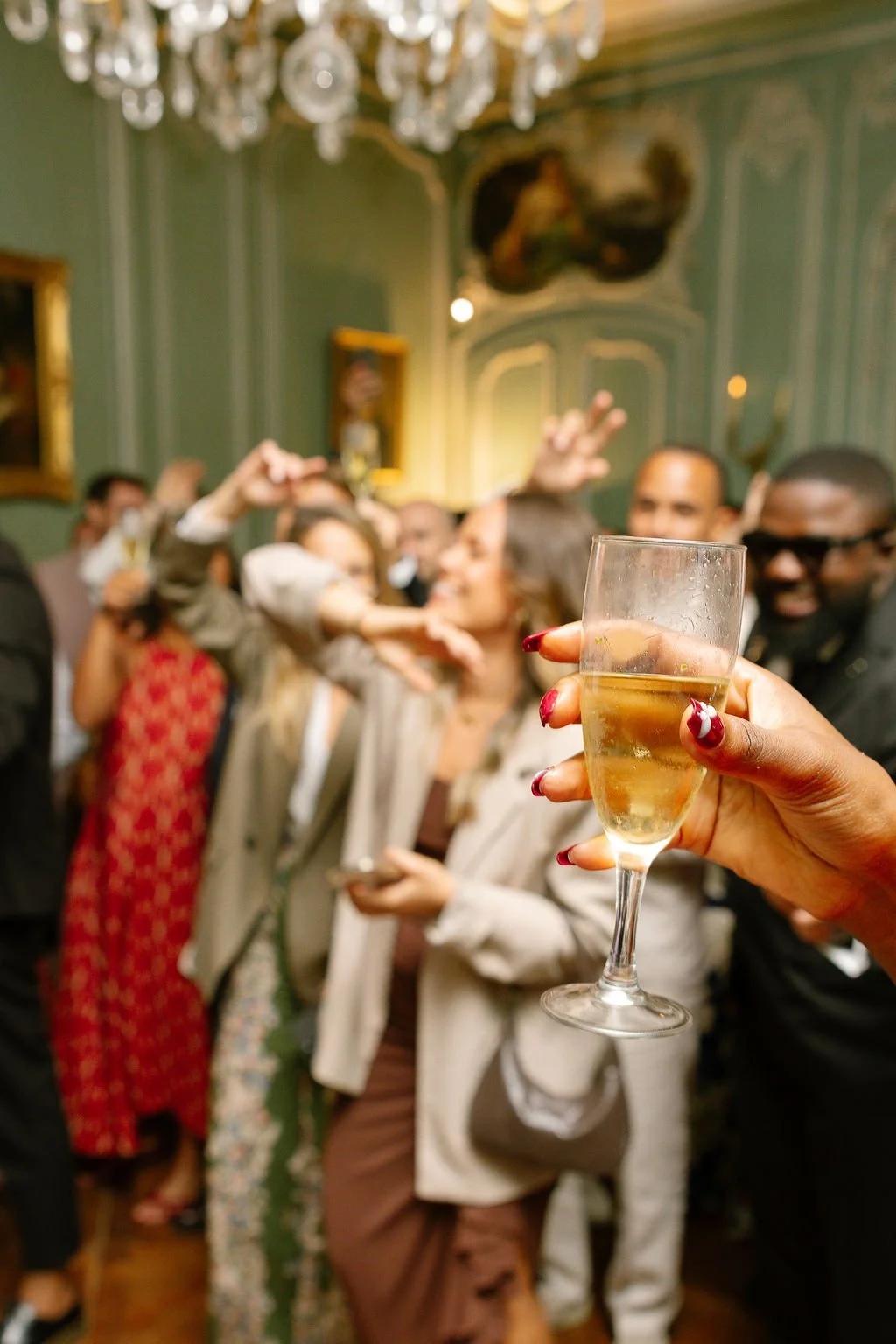 Person holding a glass of champagne at a celebration with a group of people dancing and having fun in the background.