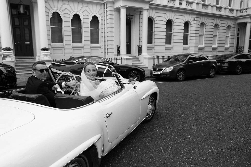 A bride and groom in a vintage car on a city street with white row houses, the bride is smiling and looking at the camera, the groom is driving and wearing sunglasses, dressed in black.