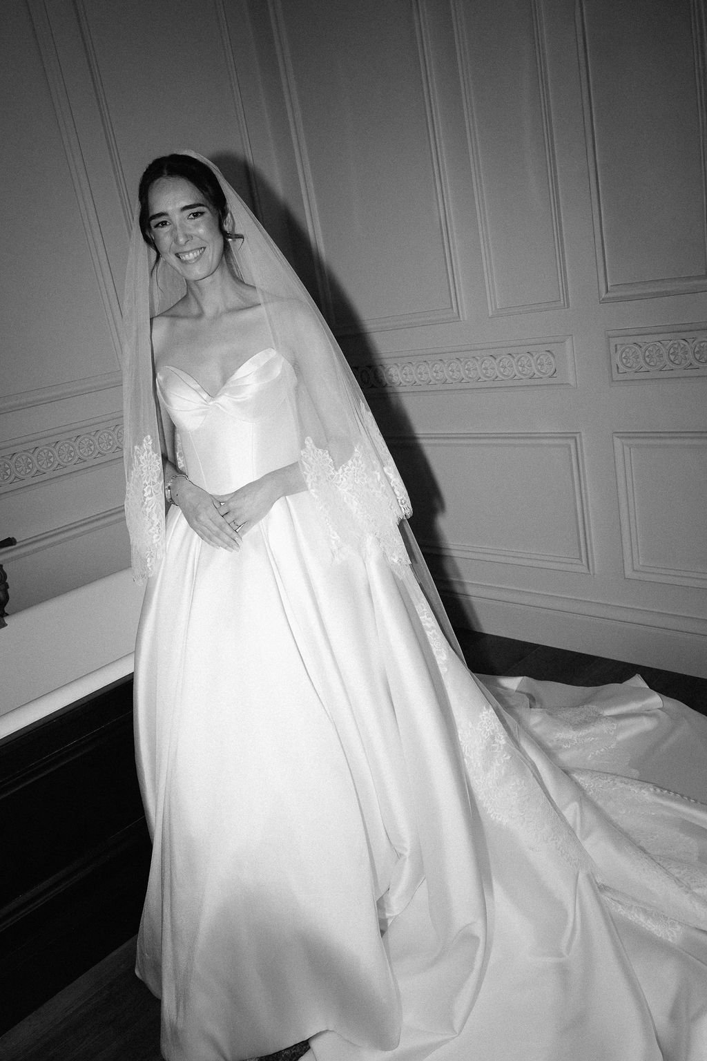 A smiling woman in a wedding dress standing in a room with patterned wall paneling, wearing a veil and a bracelet.