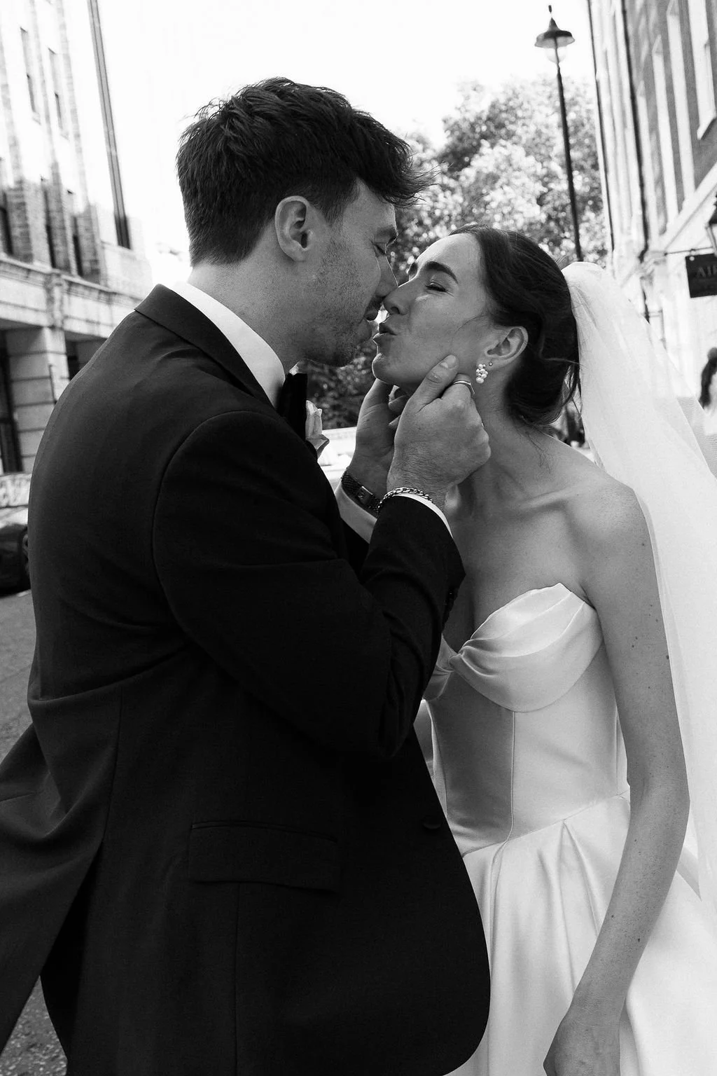 A black and white photo of a bride and groom sharing a kiss outdoors. The groom is holding the bride's chin, and they are close with their noses touching. The groom is wearing a dark suit, and the bride is in a strapless wedding gown with a veil.
