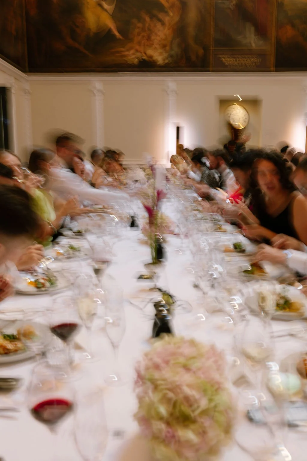 People seated at a long dining table during a formal event in a decorated room with a painting and clock on the wall