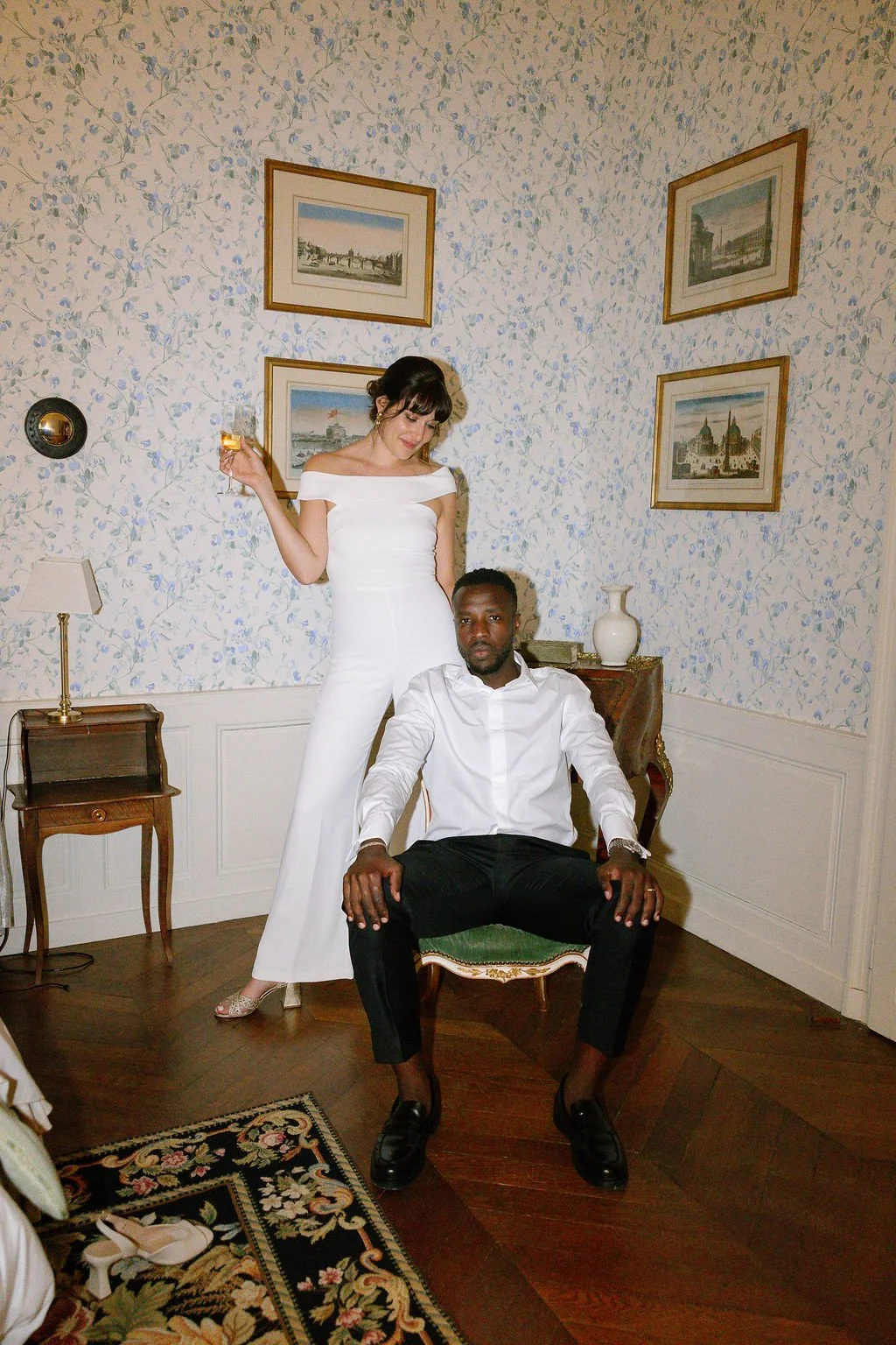A woman in a white jumpsuit stands next to a seated man in a white shirt and black pants in a decorated room with paintings, a lamp, and a carpet.