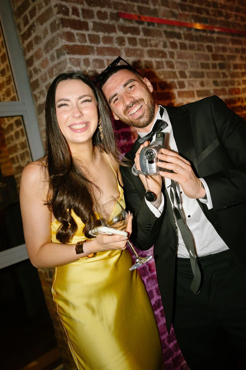 A woman in a yellow dress holding a wine glass, smiling, standing next to a man in a tuxedo holding a camera, at an event with brick walls.