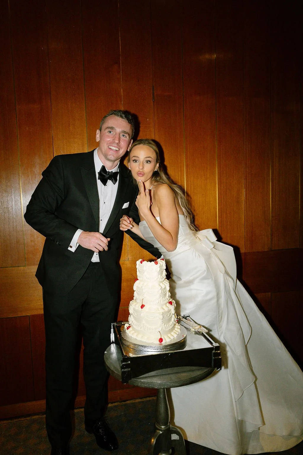 A bride and groom in wedding attire leaning over a wedding cake, making playful faces in front of a wood paneled wall.