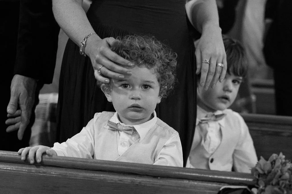 Two young boys dressed in formal attire sitting in church pews, with a woman standing behind them gently touching the first boy's head.