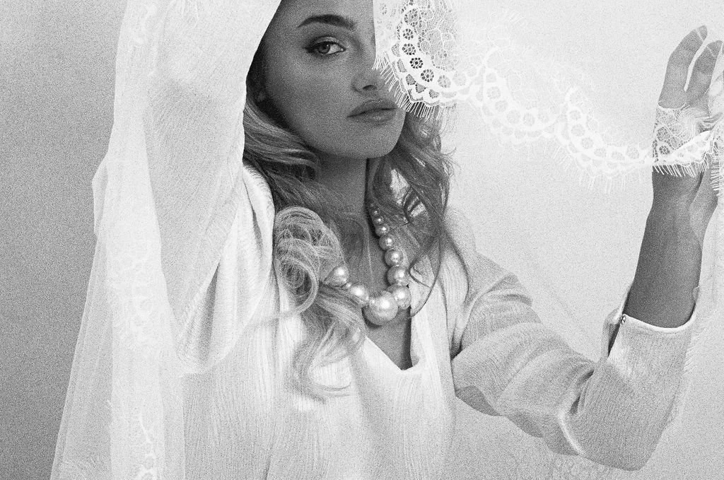 A woman with long, curly hair partially covered by lace fabric, wearing a pearl necklace and a light-colored, long-sleeved top, posing with her right hand holding the lace fabric.