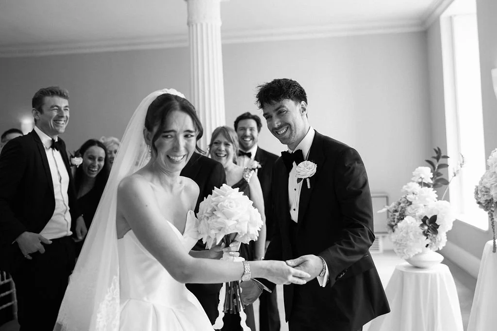 Black and white photo of a wedding ceremony with a bride and groom holding hands, smiling, surrounded by friends and family.
