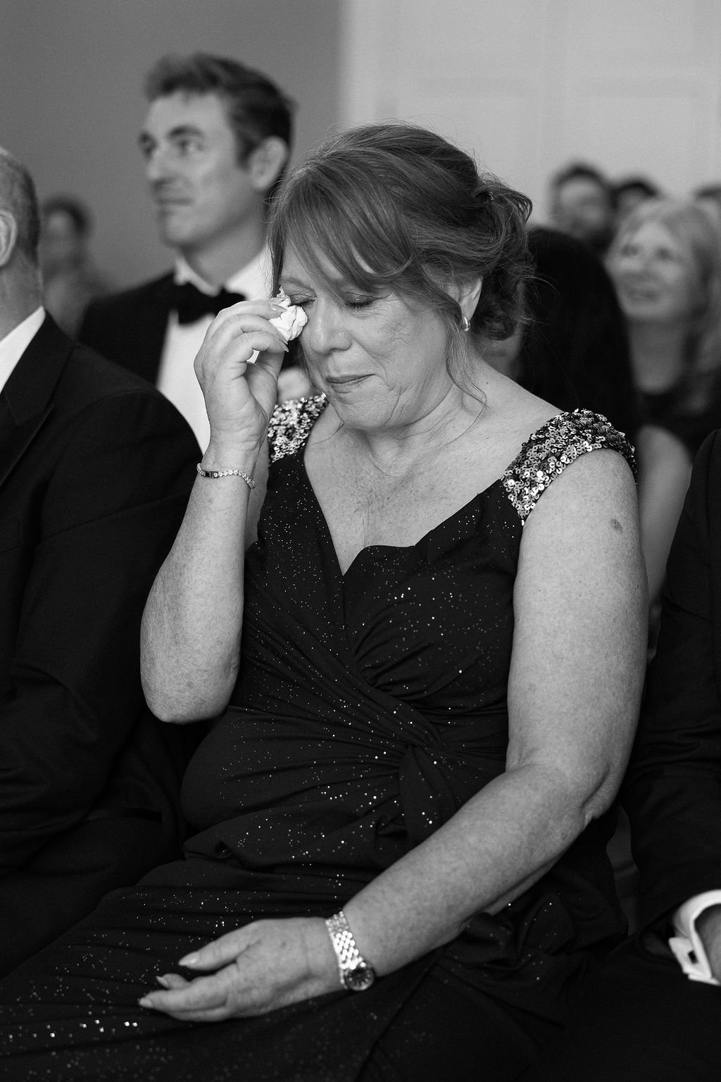 A woman in a sequined dress is wiping away tears during an event, sitting among other people dressed formally.