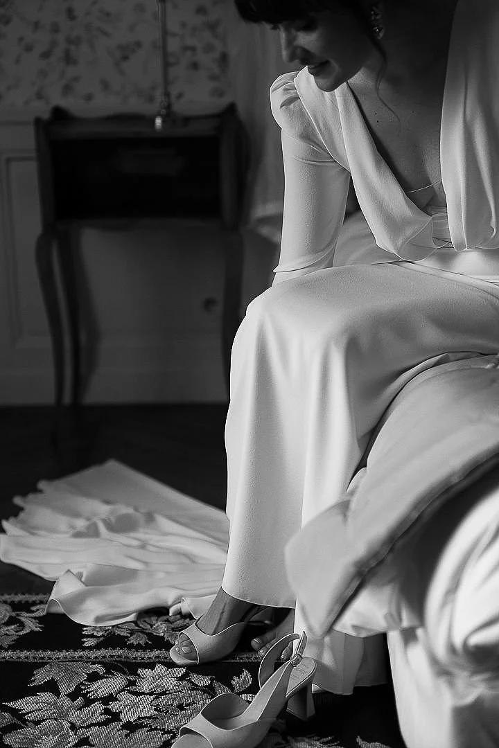 A woman in a white dress sits on a bed as she puts on high-heeled shoes in a room with vintage furniture and a floral rug.
