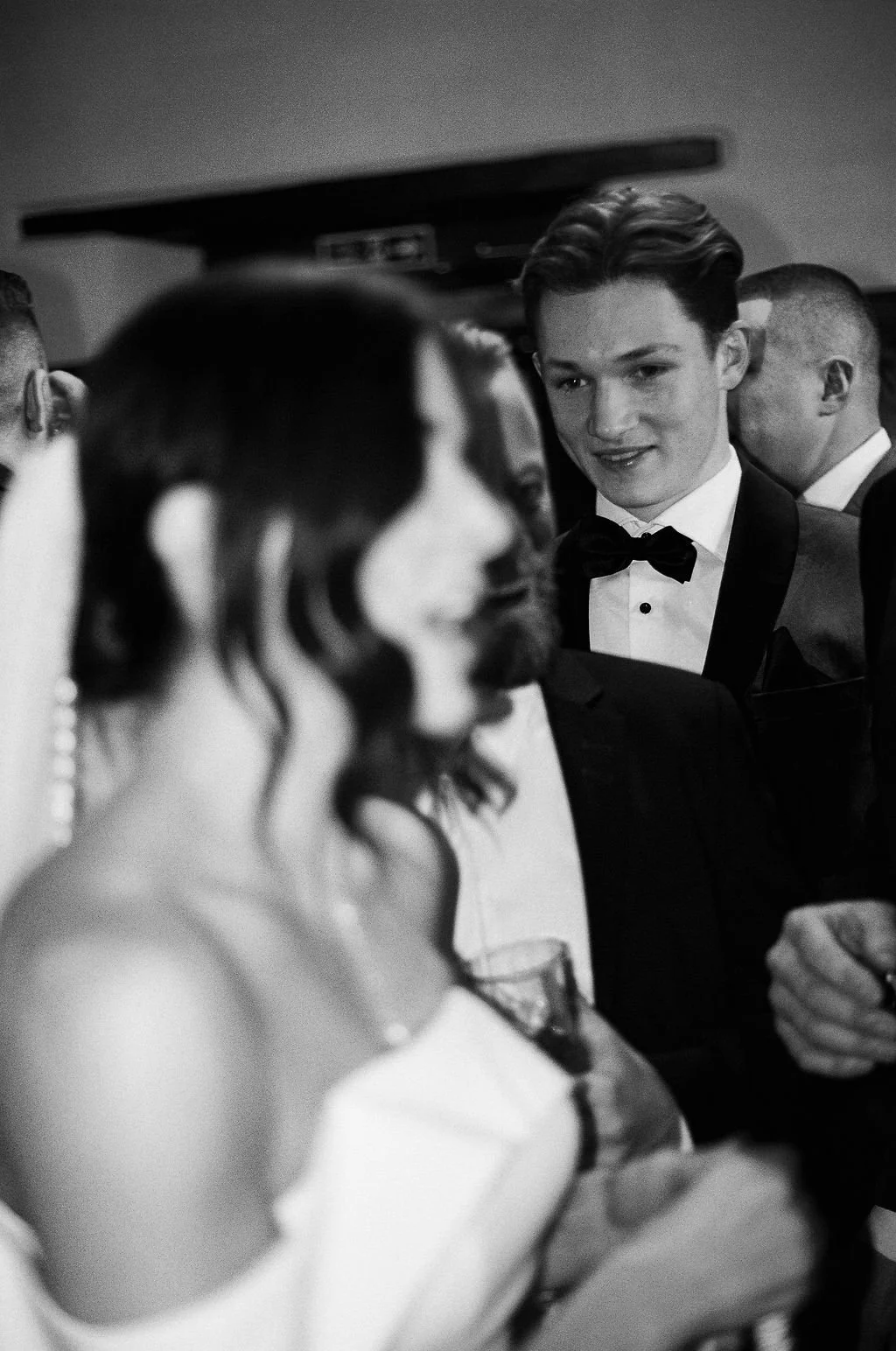 A black-and-white photo of a young man in a tuxedo and bow tie looking at a woman with curly hair and earrings, holding a drink, at a formal event or party.