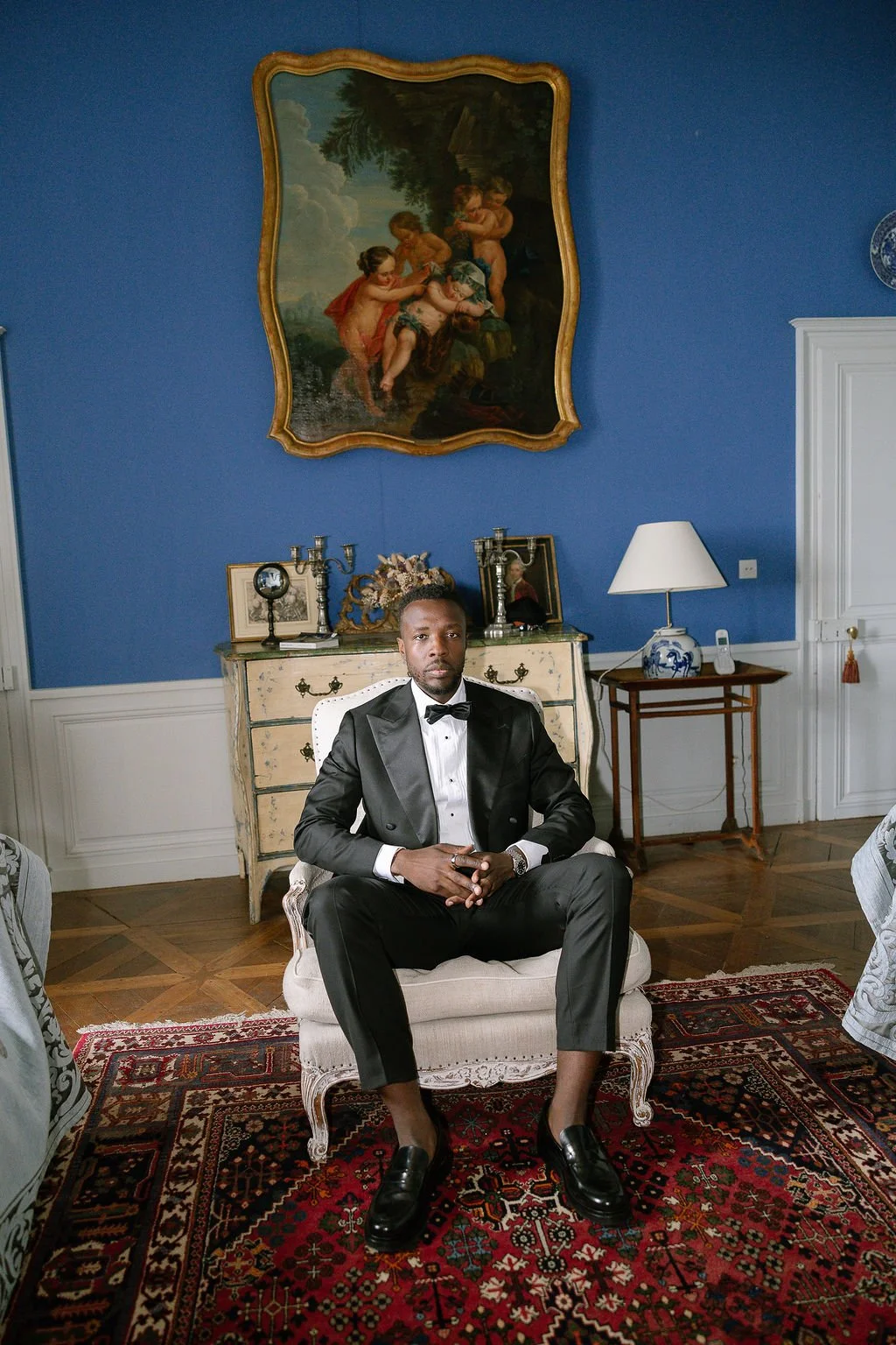 A man in a tuxedo sitting on a white chair in a luxurious room with blue walls, a vintage dresser, a lamp, and large framed paintings.