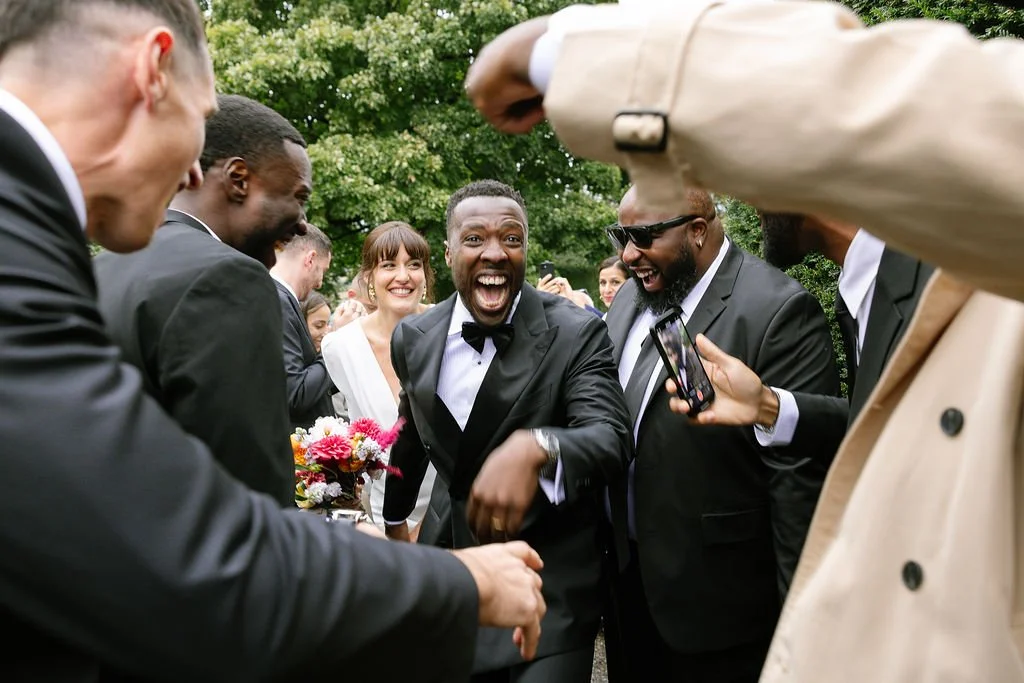 Group of people in formal attire, smiling and laughing outdoors, with one man shaking hands and others taking photos, likely at a wedding or celebration.