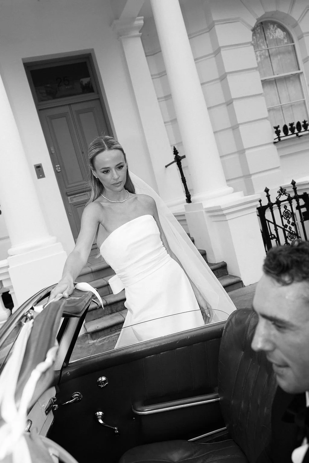 A bride in a strapless wedding dress with a veil is walking towards a vintage car, where a man in a suit is sitting, in front of a grand house with columns and a staircase.