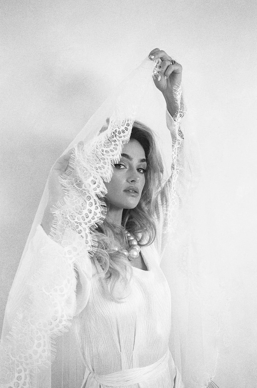 Black and white photo of a woman with long hair, wearing a translucent dress with lace sleeves, holding a lace veil above her head.