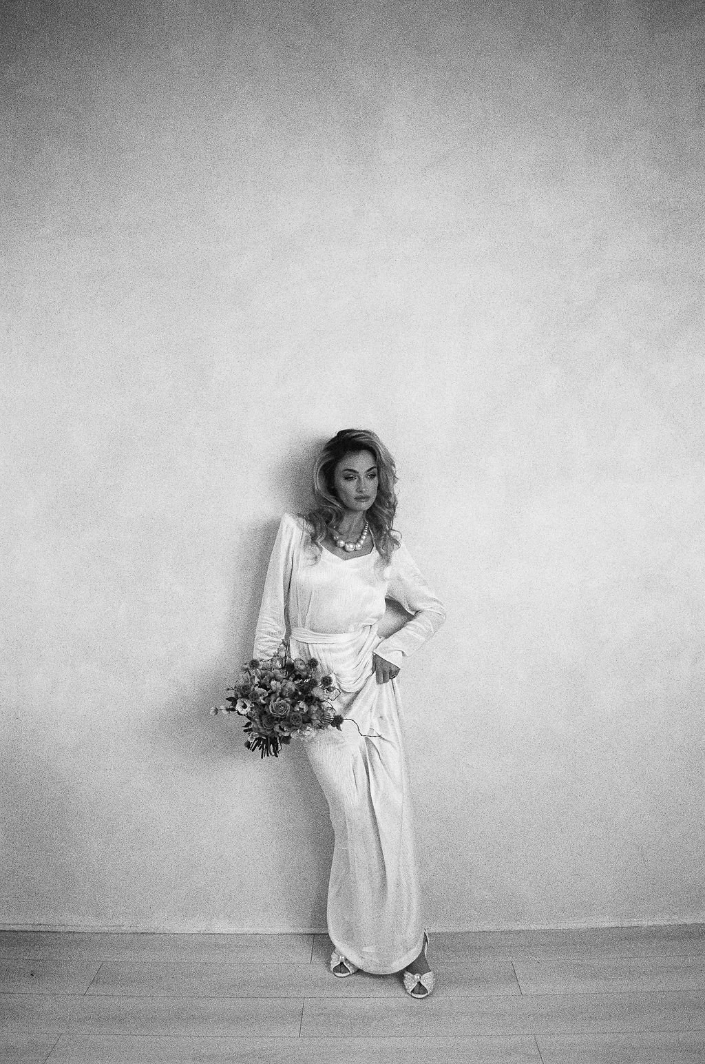 Black and white photo of a woman in a wedding dress holding a bouquet of flowers, standing against a plain wall with one hand on her hip.