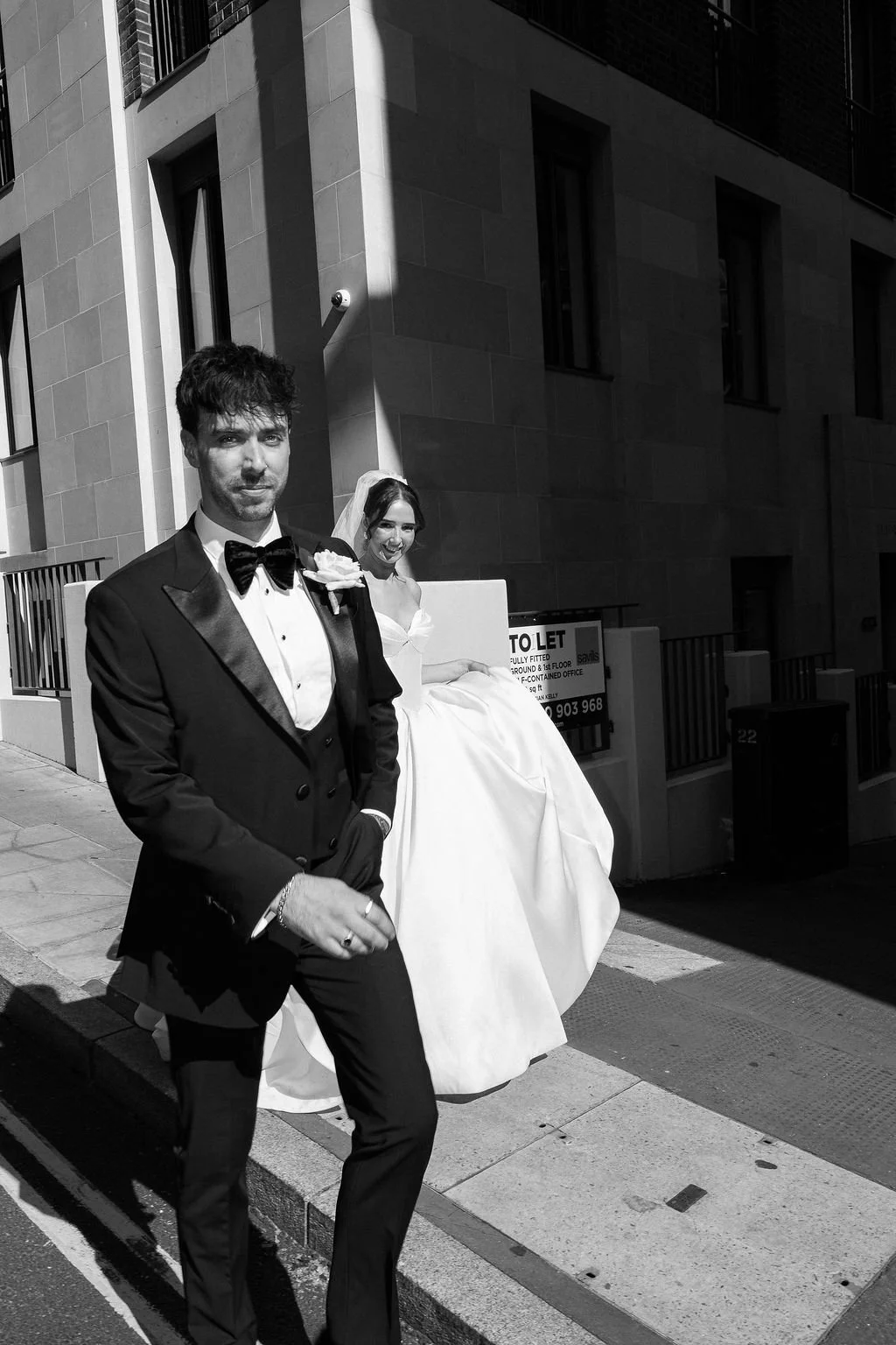 Black and white photo of a man in a tuxedo and a woman in a wedding dress walking on a city sidewalk.
