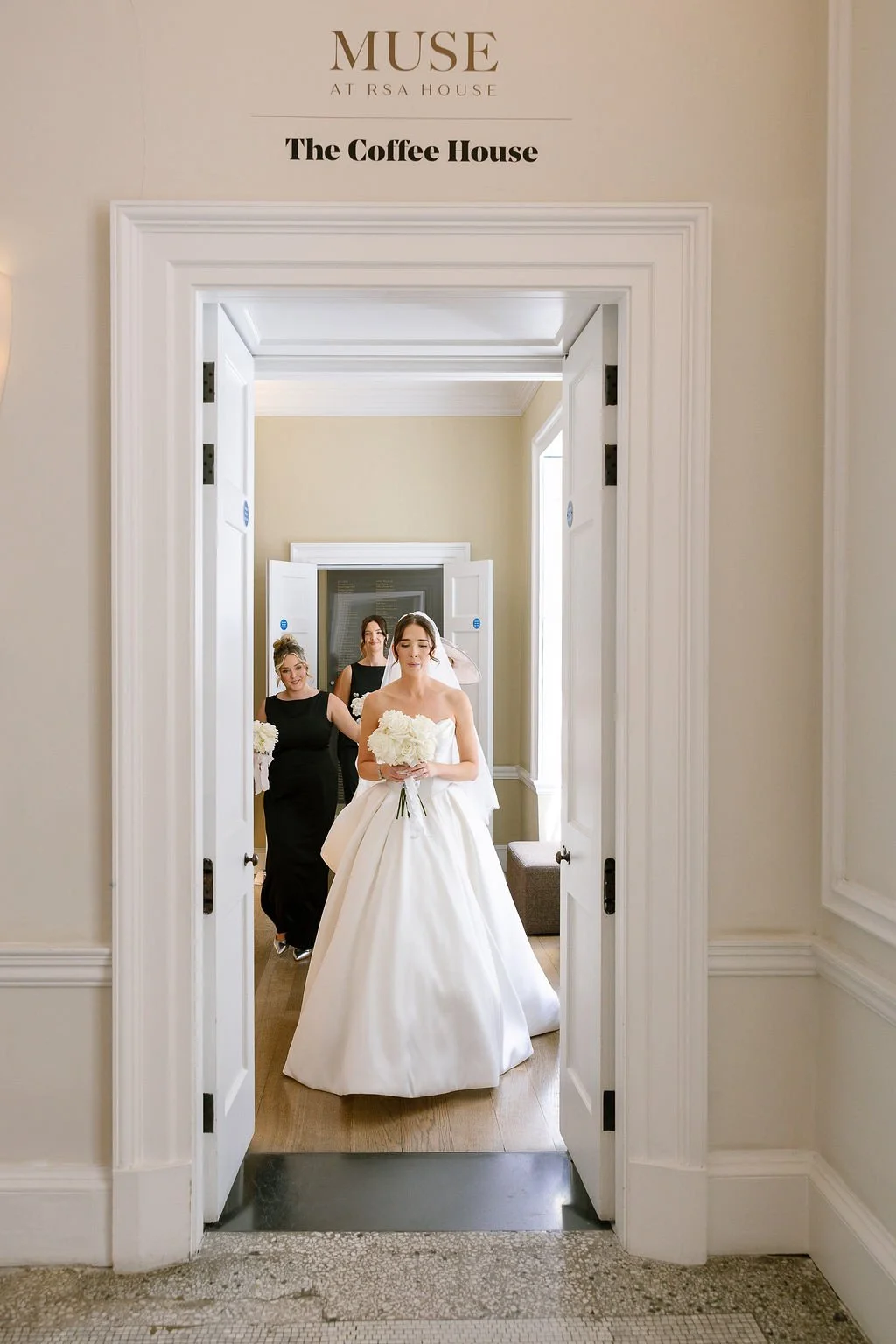 A bride in a white wedding gown holding a bouquet walking into a room with three women, two dressed in black, following her, with a sign above the door reading 'Muse at RSA House The Coffee House'.
