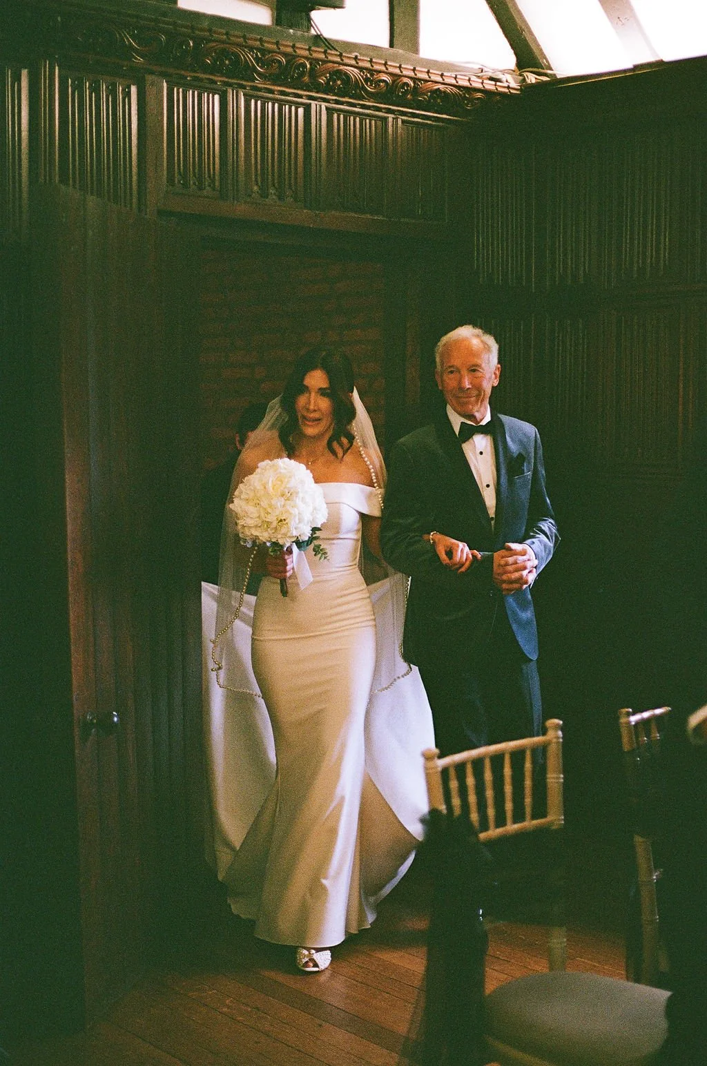 A bride in a white wedding gown holding a bouquet of flowers, walking down the aisle with an older man in a tuxedo, in a wood-paneled room.