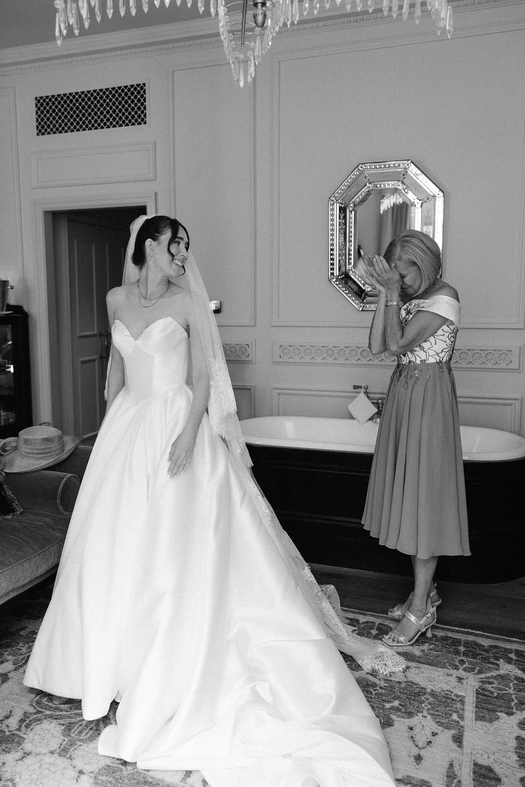 A bride in a wedding dress laughing while a woman, possibly her mother, covers her face with her hands in a moment of emotion in a decorated room.
