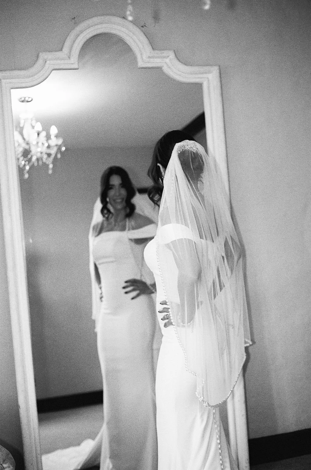 A bride in a wedding dress and veil looking at herself in a mirror, with her reflection showing her smiling.