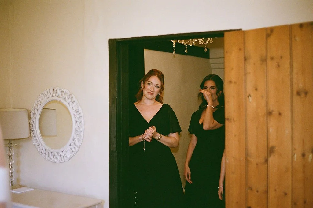 Two women standing inside a doorway, both wearing black dresses, with one smiling and the other covering her mouth as if laughing.